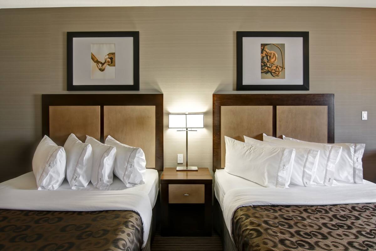 Habitación de hotel con dos camas, cada una con ropa blanca y colchas estampadas. Hay cuadros enmarcados sobre las camas, una lámpara de noche y un pequeño cajón entre ellas.