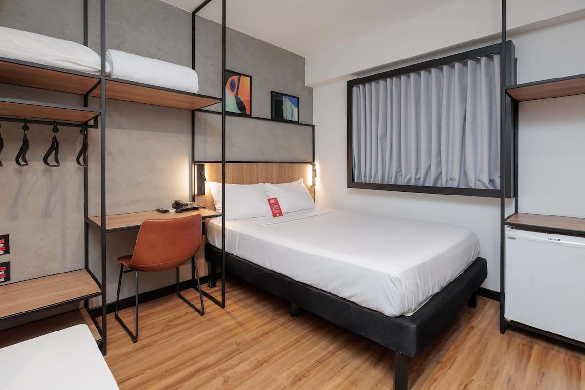 A compact hotel room with a bunk bed, a desk with a chair, a small kitchenette, and a window with curtains.