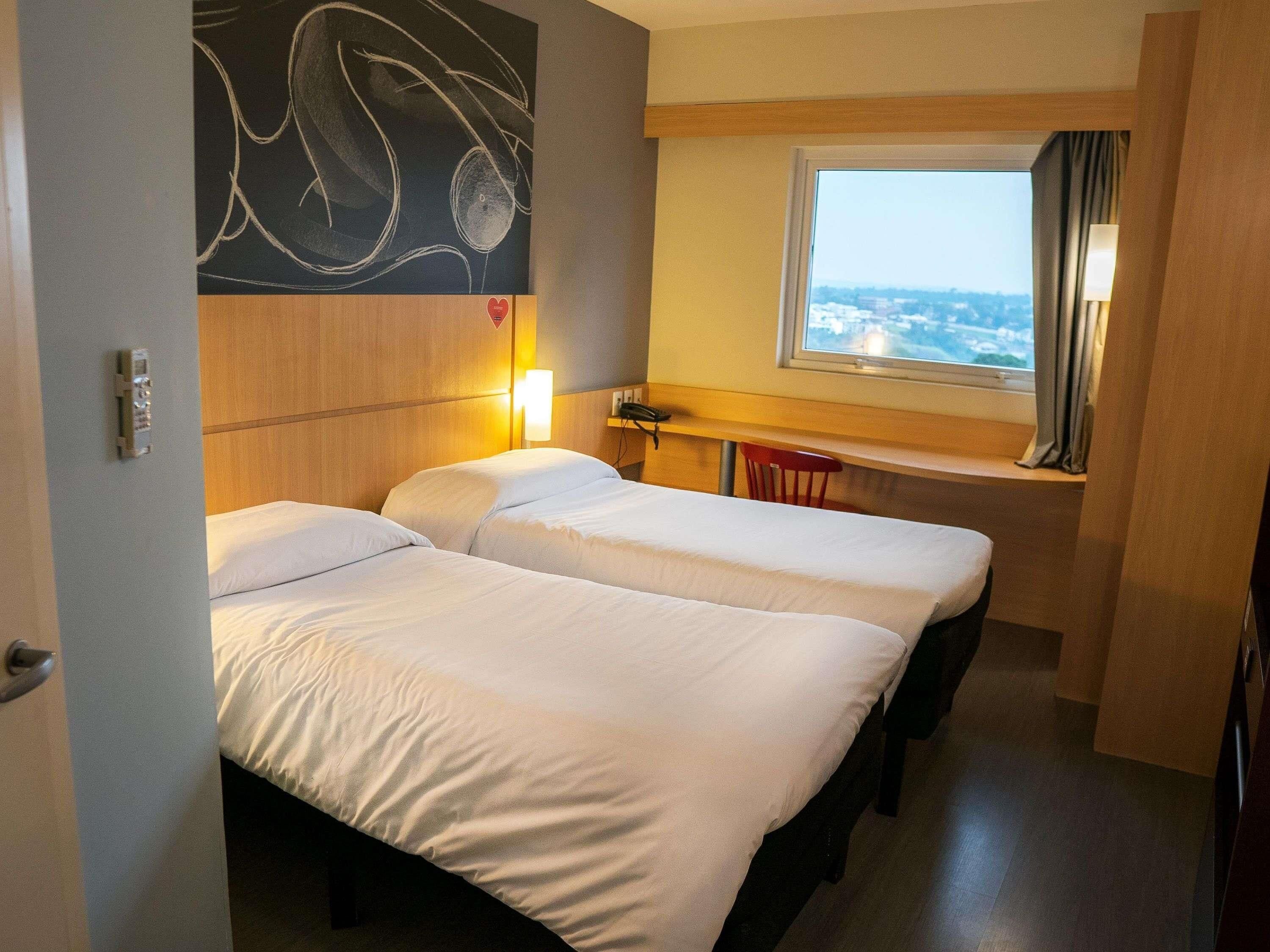 A hotel room with two beds, a window with a view, a desk with a lamp, and a wall art piece.