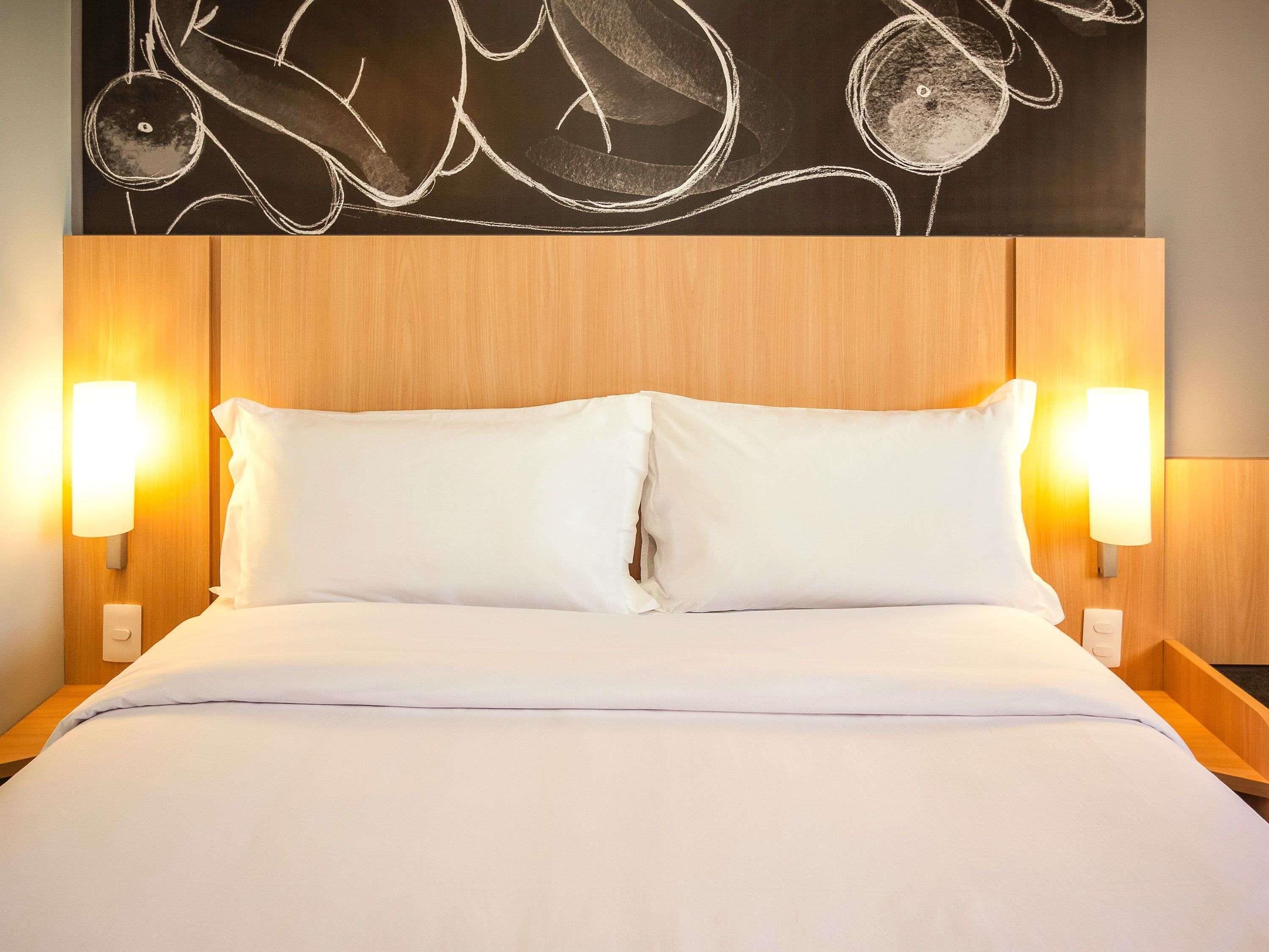 A hotel room with a wooden headboard, two white pillows, a white bedspread, and wall-mounted lamps.