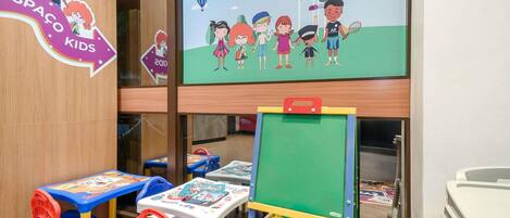 Un'area giochi per bambini con tavoli e sedie colorate, un grande schermo televisivo che mostra personaggi dei cartoni animati e un cavalletto verde.