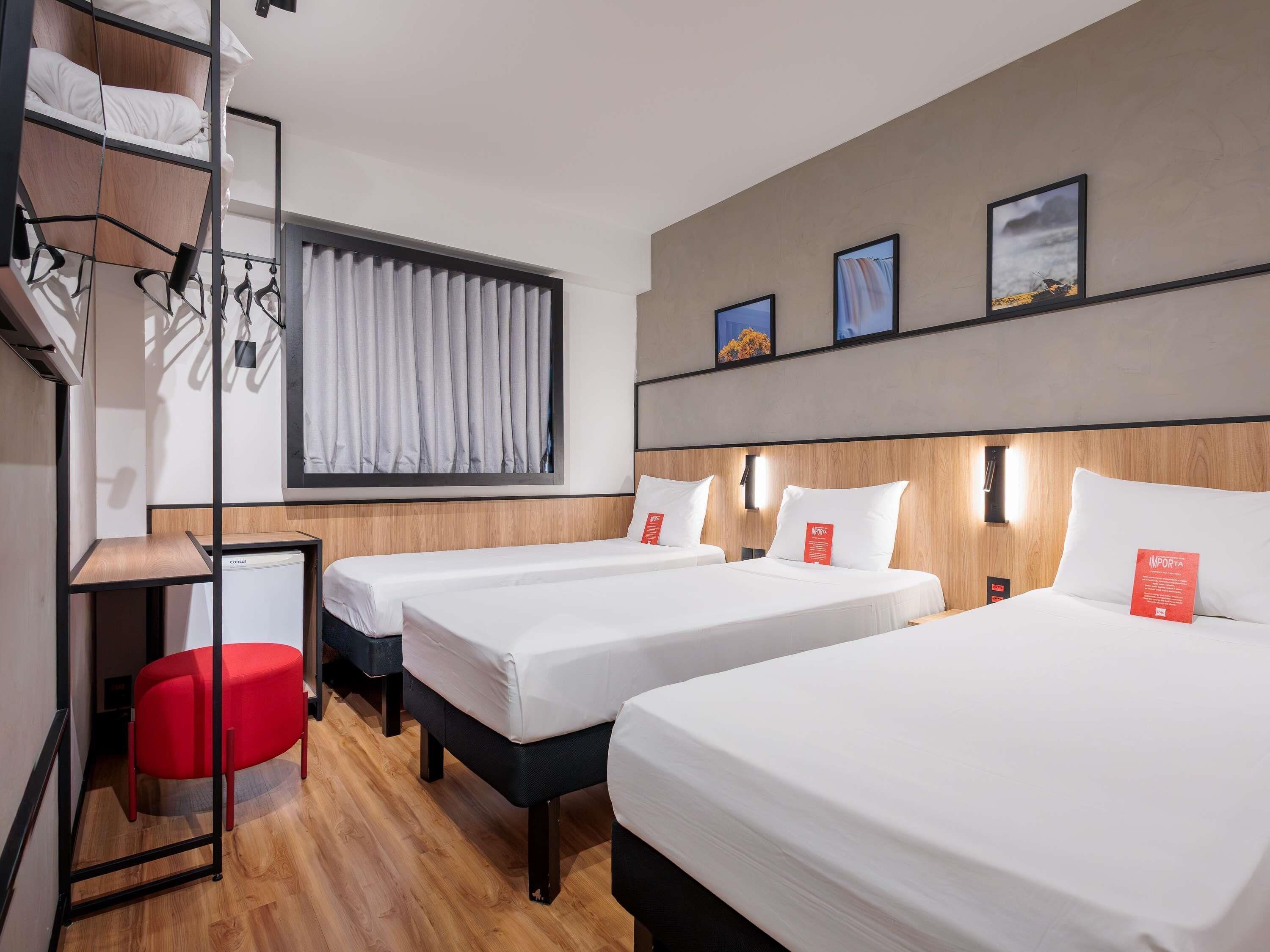 A hotel room with two beds, a red chair, a desk, and a window with curtains.
