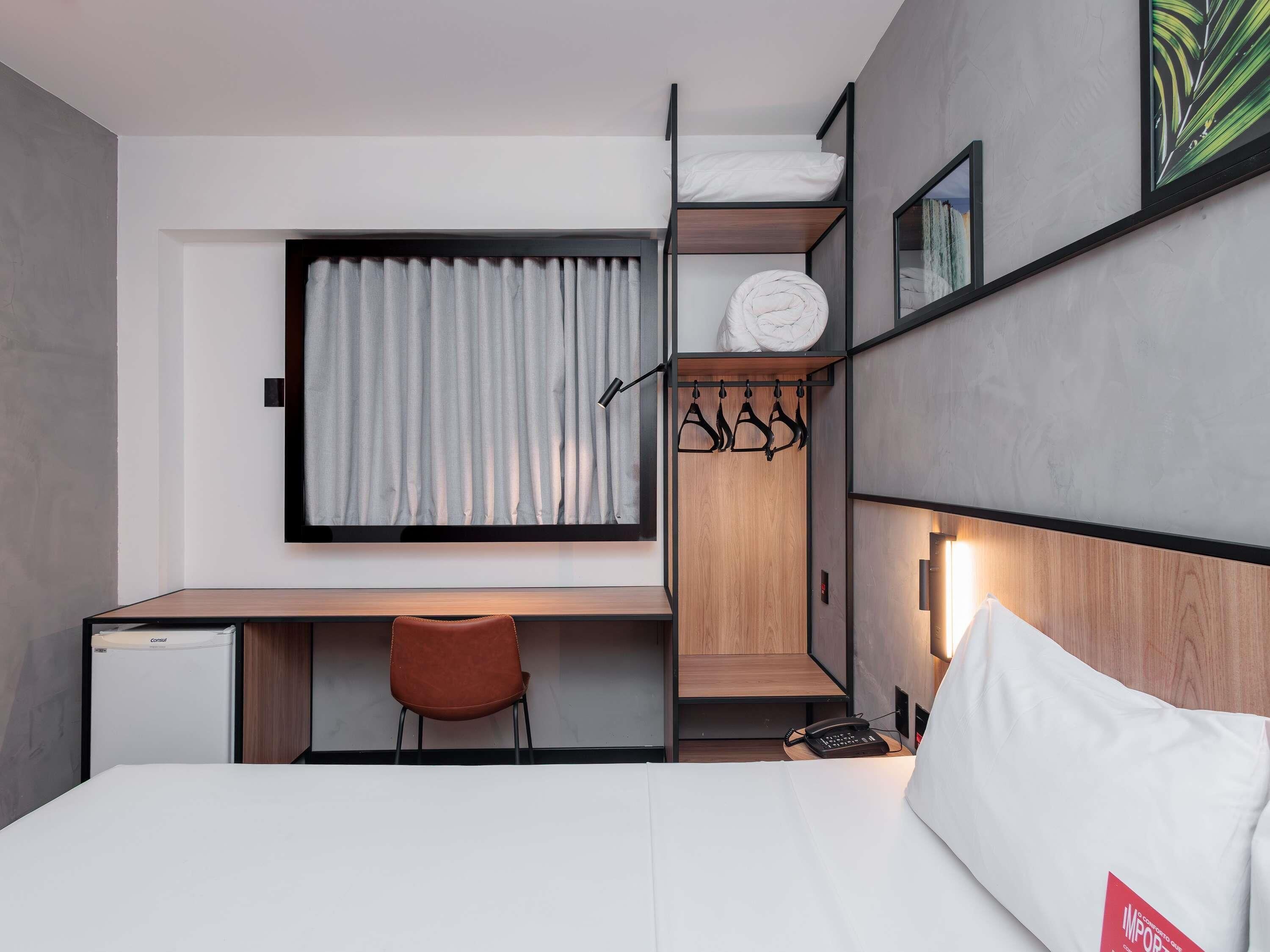 Una habitación de hotel moderna con cama, escritorio, silla, un refrigerador pequeño, armario y ventana con cortinas.