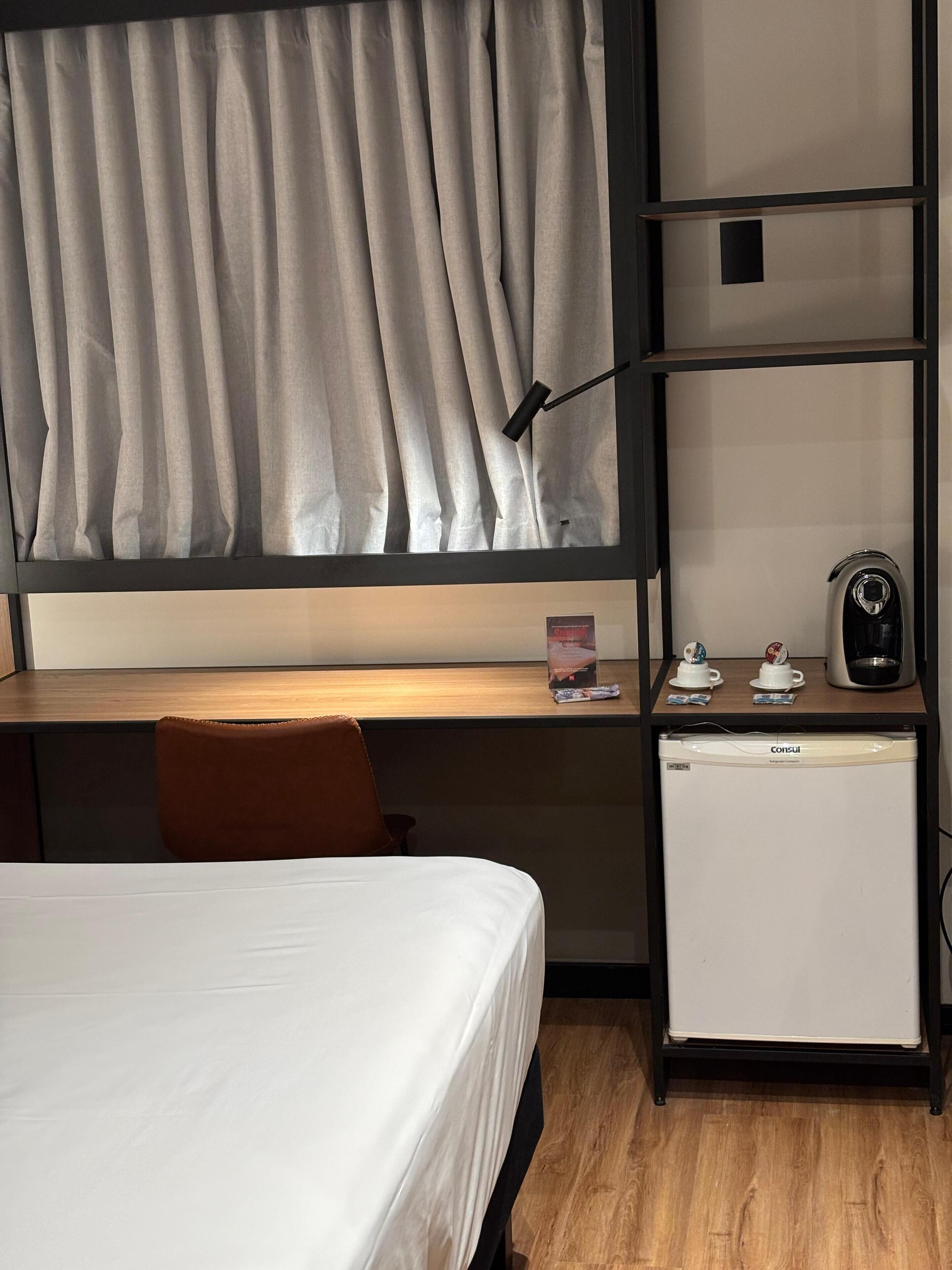 A compact hotel room with a bed, a desk with a computer, a small refrigerator, and a window with curtains.