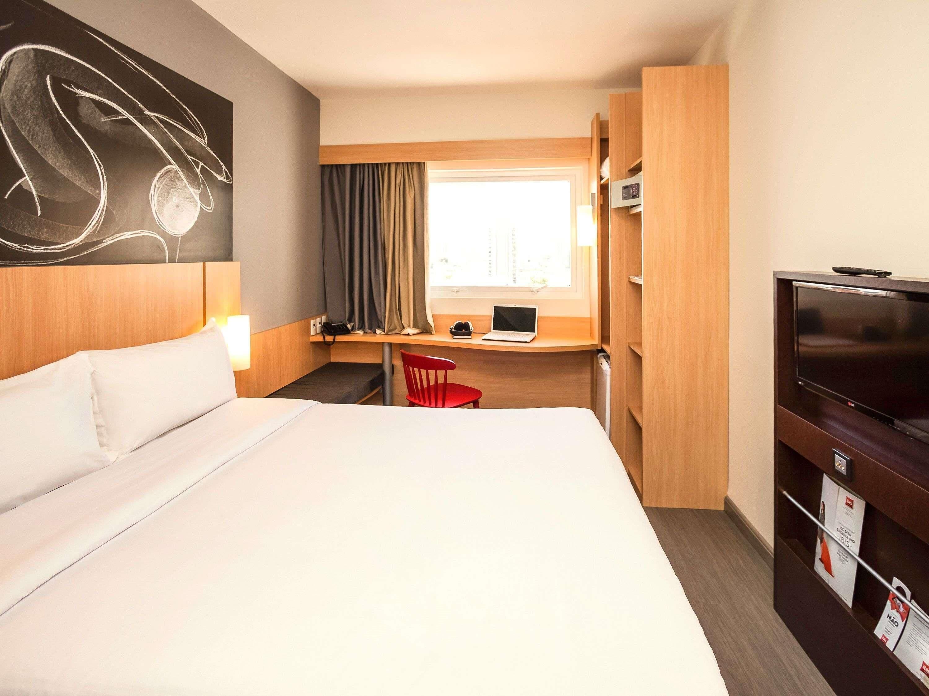 Quarto de hotel com uma cama grande, uma escrivaninha com um laptop, uma televisão e uma janela com cortinas.