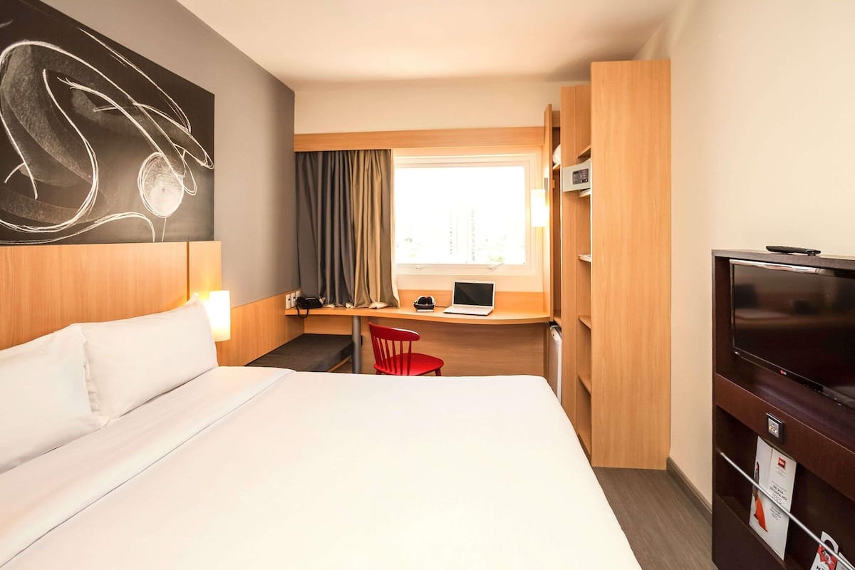 A hotel room with a large bed, a desk with a laptop, a television, and a window with curtains.