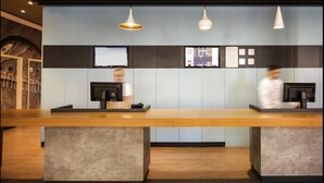 A reception area with a long wooden counter, two receptionists, and a wall-mounted TV. - ibis Foz do Iguaçu (Foz do Iguaçu)