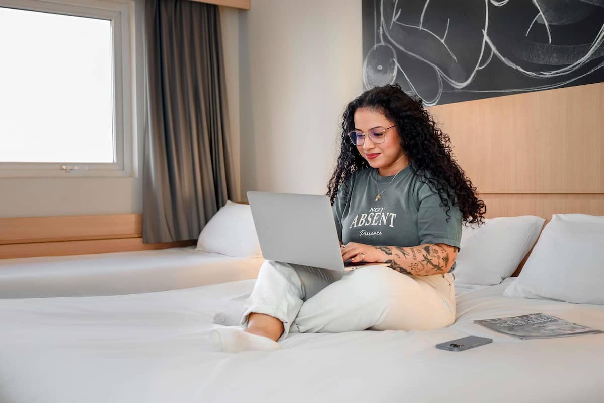 A person sitting on a bed with a laptop, using a smartphone, and a magazine on the bed.