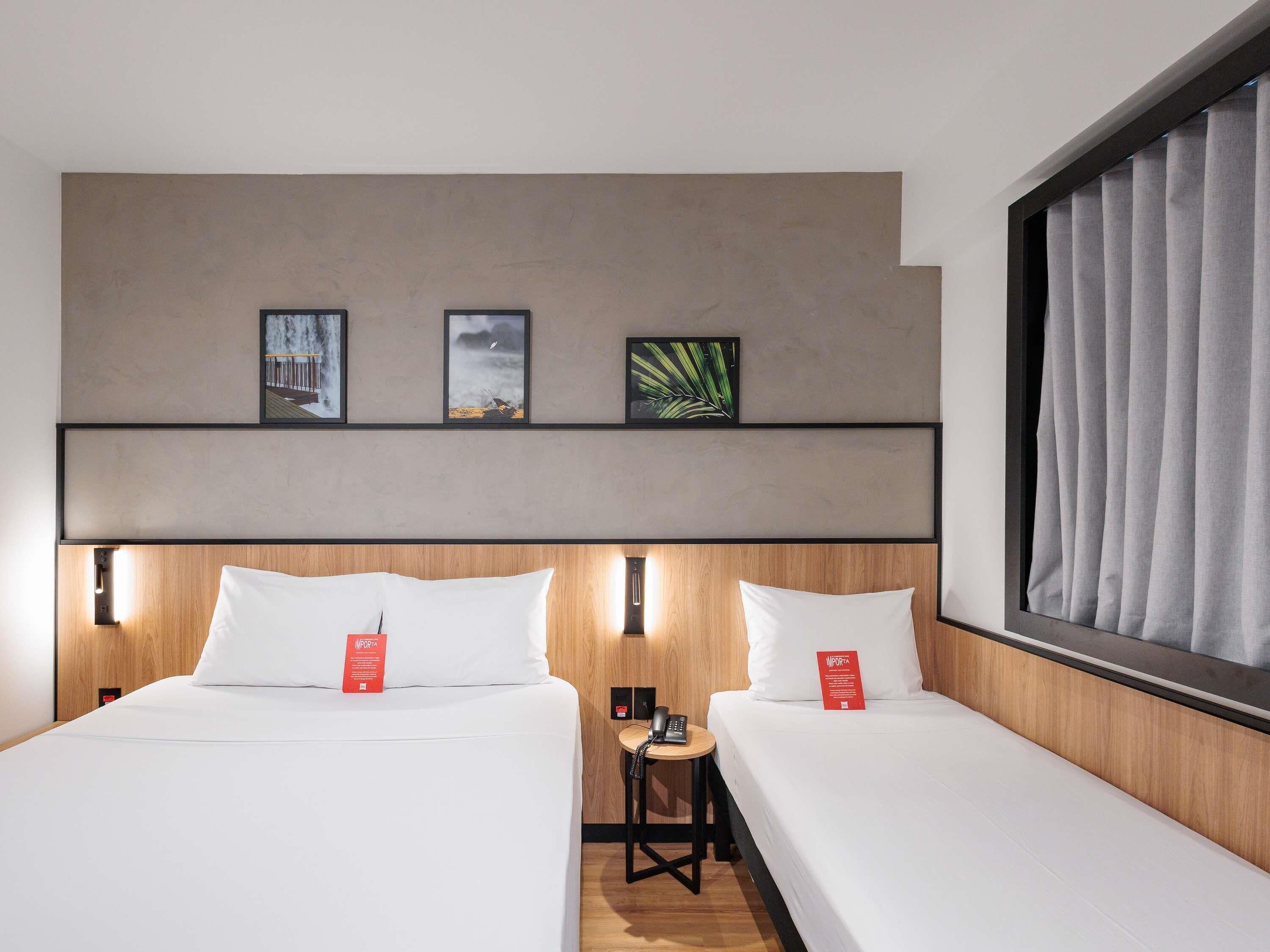 A hotel room with two beds, a wooden headboard, a small table with a phone, and framed pictures on the wall.