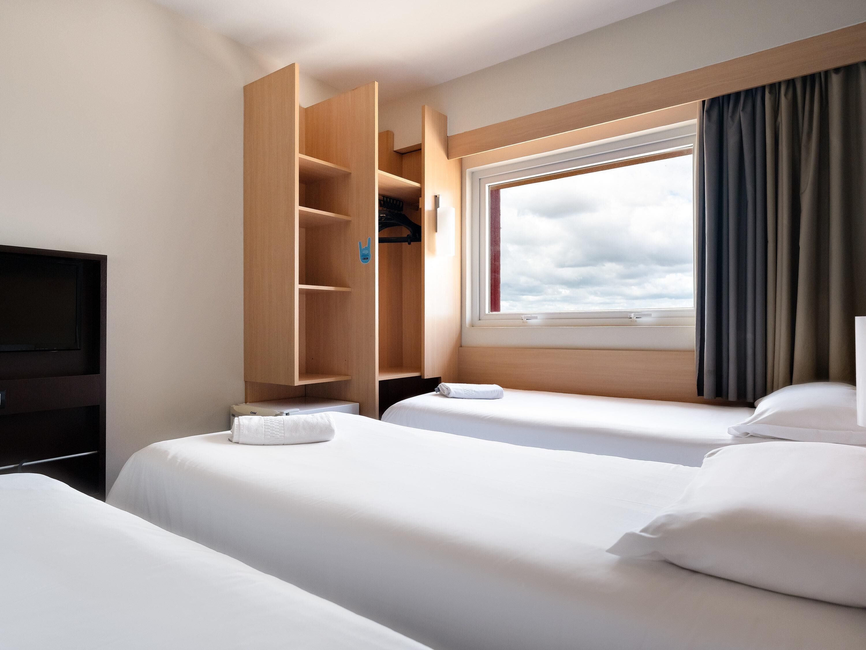 A hotel room with two beds, a TV, a wooden wardrobe, and a window with curtains.