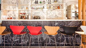 A bar area with a modern design, featuring a counter with shelves displaying various bottles and glasses, and a wall with abstract line art. - ibis Foz do Iguaçu (Foz do Iguaçu)