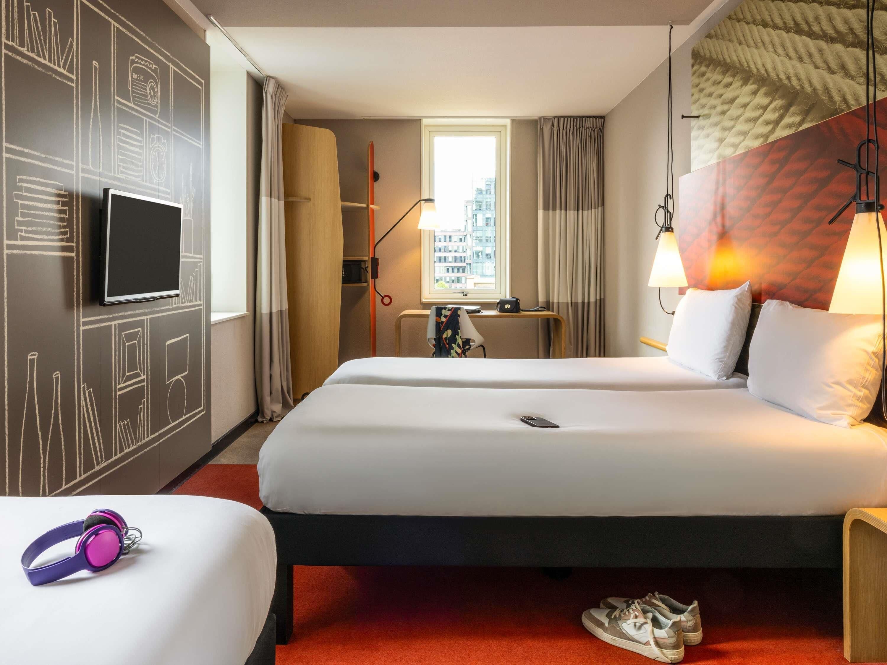 A hotel room with two beds, a TV, a desk with a lamp, and a view of the city through the window.