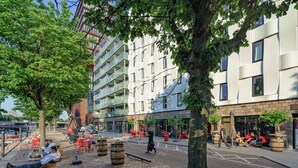 Une rue urbaine moderne avec des terrasses, des arbres et un immeuble à plusieurs étages doté de balcons.