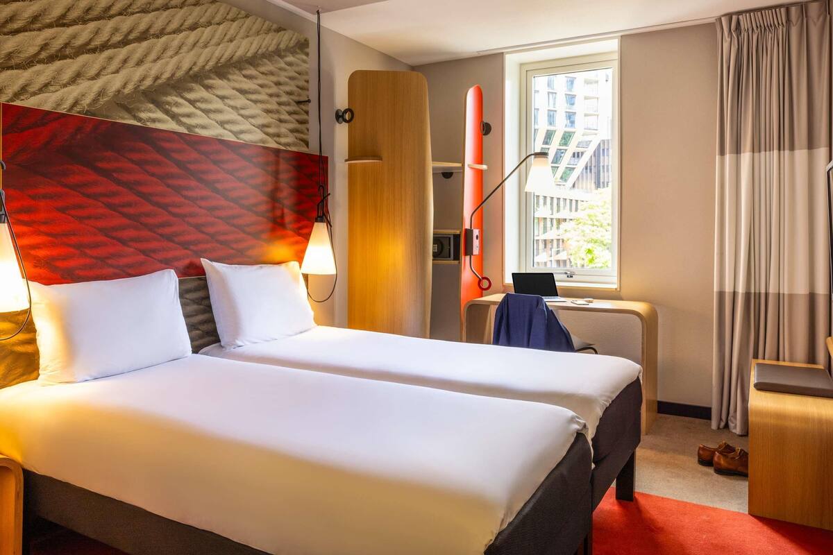 A hotel room with two beds, a desk, a window with a view of buildings, and a red carpet.