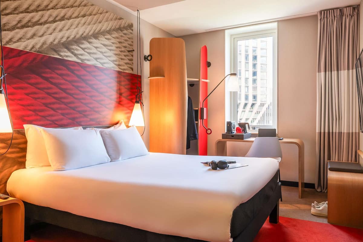 A hotel room with a bed, a desk, a chair, a window, and a red wall.