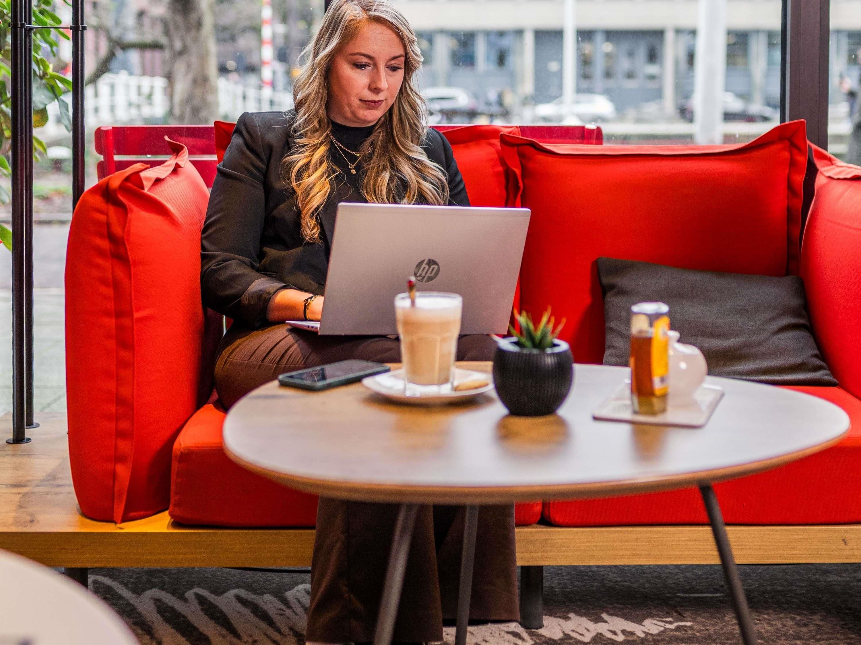 A person sitting on a red sofa with a laptop, a smartphone, and a beverage on a table.