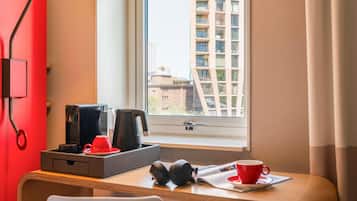Habitación de hotel con escritorio, cafetera, taza de café, auriculares y ventana con vista a edificios.
