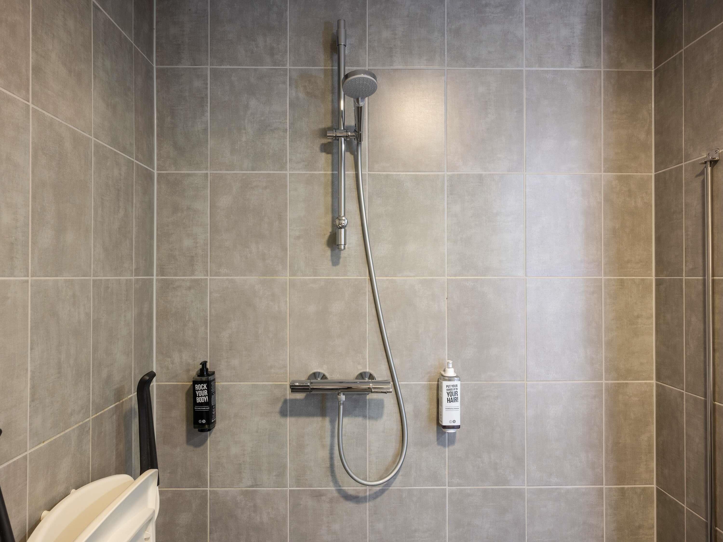 A shower with a handheld showerhead and a wall-mounted faucet, a soap dispenser, and a black bottle of shampoo.