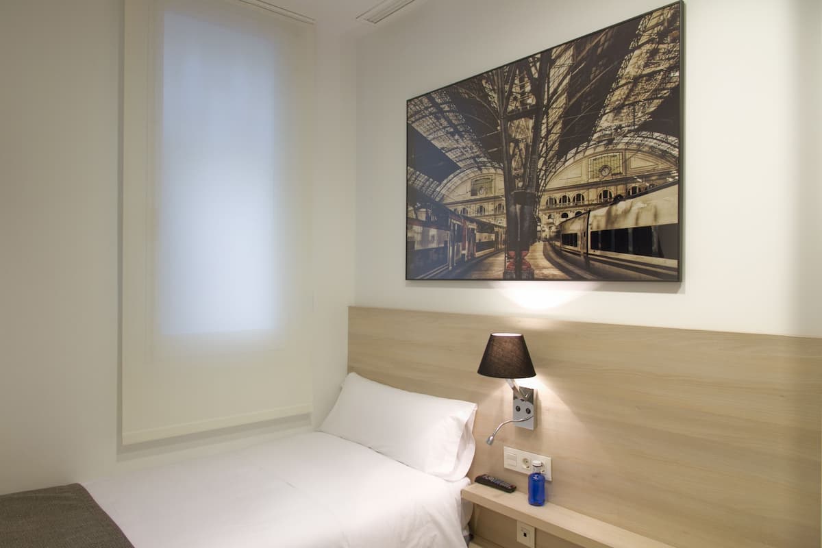 A hotel room with a bed, a nightstand, a lamp, and a wall-mounted painting of a train station.