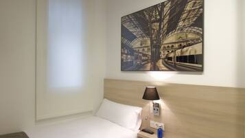 Habitación de hotel con cama, mesita de noche, lámpara y un cuadro colgado en la pared que representa una estación de tren.