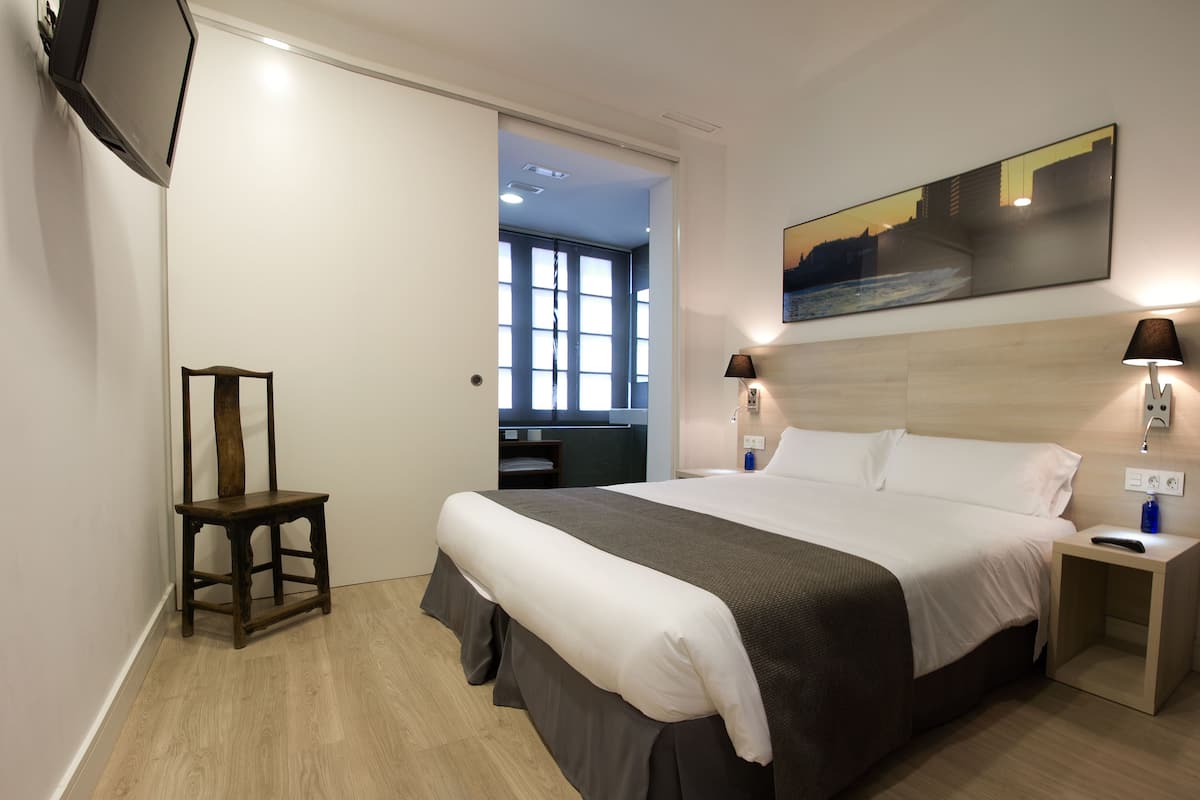 A modern hotel room with a large bed, two bedside tables, a TV mounted on the wall, and a painting above the bed.