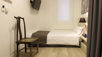 A small, neatly arranged bedroom with a single bed, a chair, a bedside table with a lamp, a flat-screen TV mounted on the wall, and a window with a view of a building.