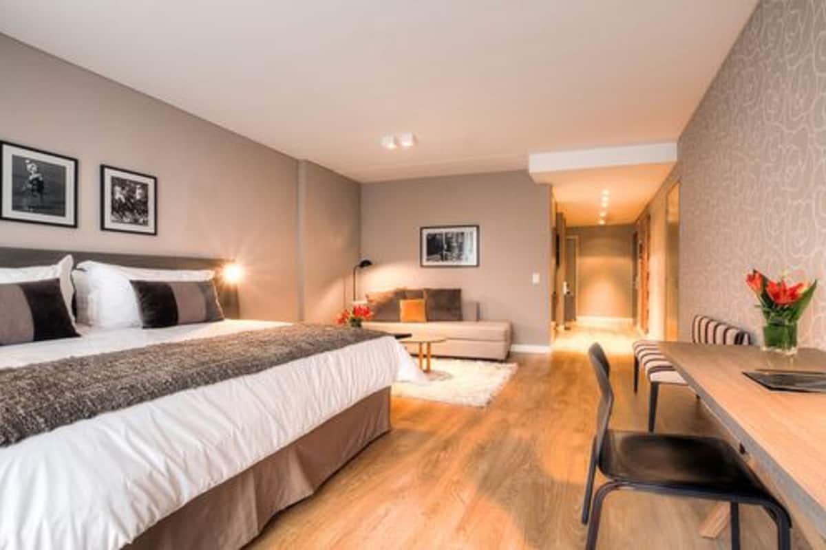 A modern bedroom with a large bed, a sofa, a dining table with chairs, and a hallway leading to another room.