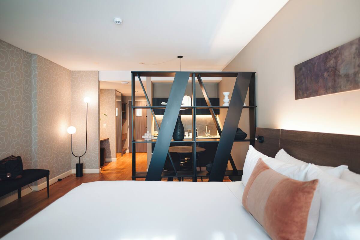 A hotel room with a bed, a desk, and a chair. There is a lamp and a painting on the wall.