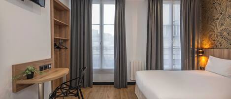 A hotel room with a bed, a desk with a plant, a chair, a TV mounted on the wall, and a window with curtains.
