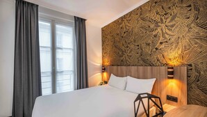 A hotel room with a large bed, grey curtains, a patterned wall, and a nightstand with a lamp. - Hôtel Daval (Paris)