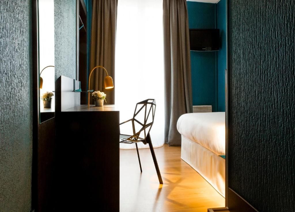 Una habitación de hotel con una cama, un escritorio con lámpara, una silla y un ventanal con cortinas.
