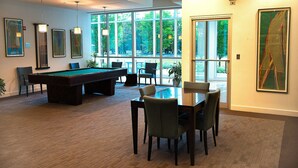 A room with a pool table, chairs, and a dining table with chairs. There are large windows and artwork on the walls. - Crystal Quarters Furnished Apartments- Concord (Arlington)