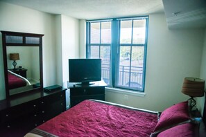 Een slaapkamer met een bed, een dressoir, een televisie, een raam met jaloezieën en een lamp.