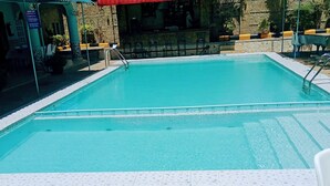 A rectangular swimming pool with a tiled edge, a stone deck in the foreground, and a bar area with tables and chairs in the background.