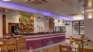 A bar area with a purple counter, multiple taps, and a variety of bottles. Tables with chairs and wine glasses are set up for dining.