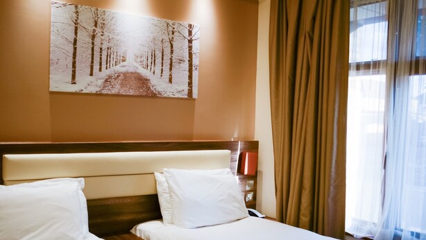 A hotel room with two beds, a painting of a snowy path on the wall, and a window with curtains.