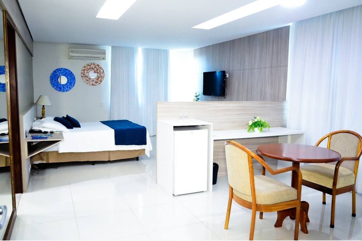 A modern hotel room with a bed, bedside table, lamp, TV, and a small desk with a chair.
