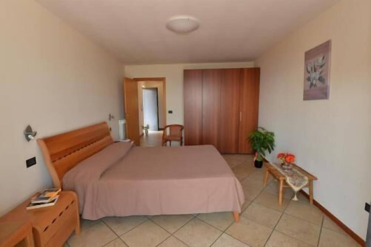 A bedroom with a bed, a wooden nightstand, a chair, a wooden wardrobe, a small table with a plant, and a door leading to another room.