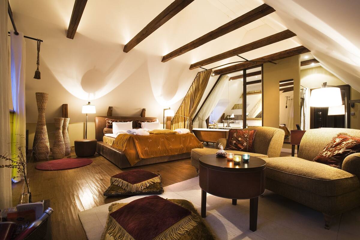 A cozy attic bedroom with a large bed, a sofa, and a small table. There are wicker baskets, a lamp, and a mirror on the walls.