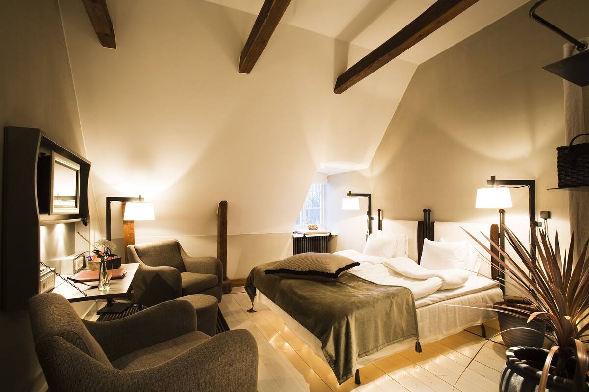 A cozy, well-lit bedroom with a bed, two armchairs, a desk with a lamp, and a potted plant.