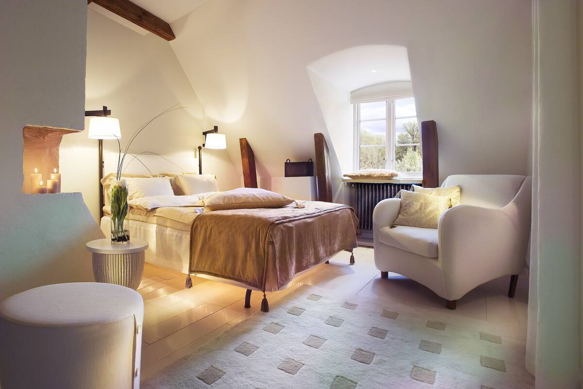 A cozy bedroom with a bed, bedside table, lamp, and a window with a view.