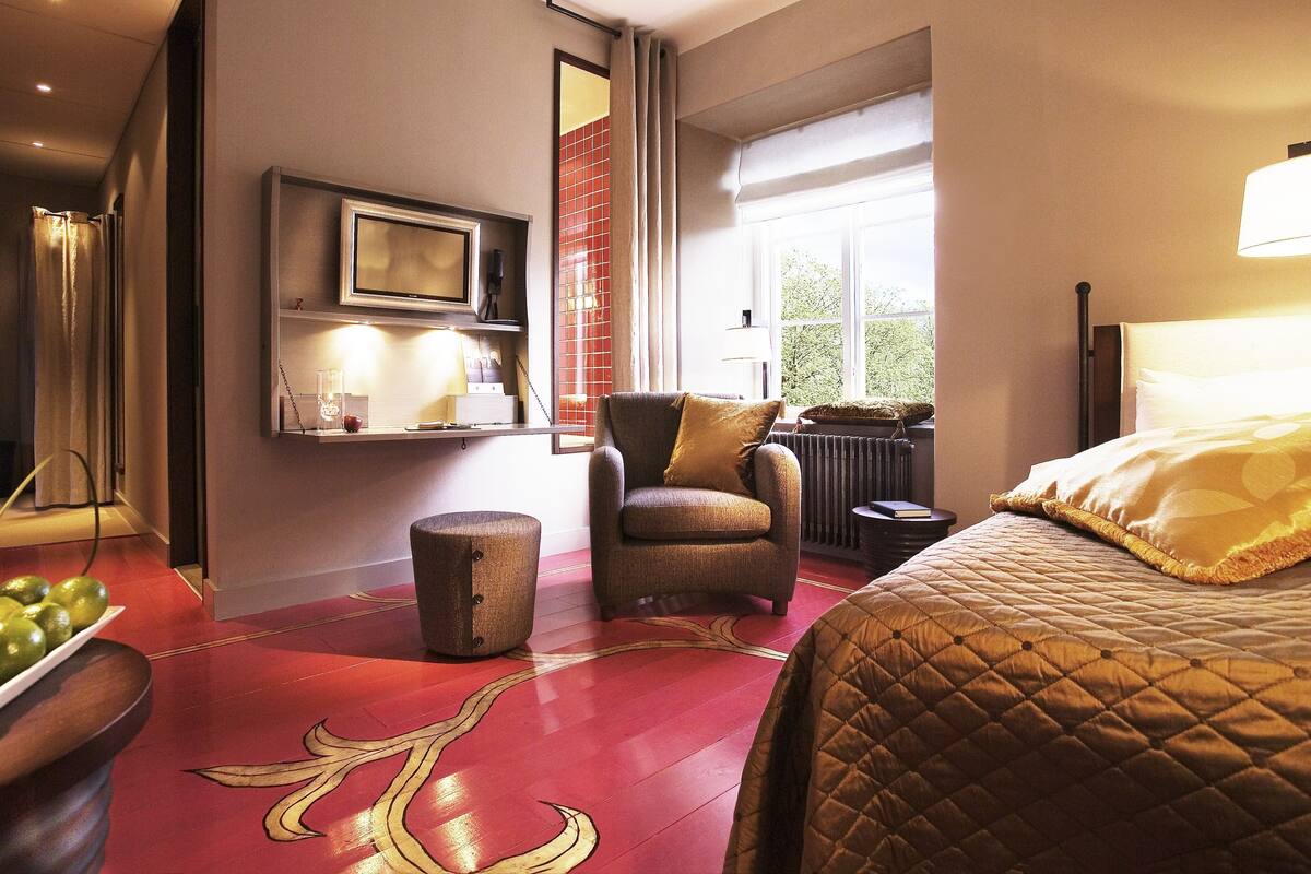 A hotel room with a red floor, a bed with a quilted cover, a chair, a small table with a book, a shelf with decorative items, and a window with a view of greenery.