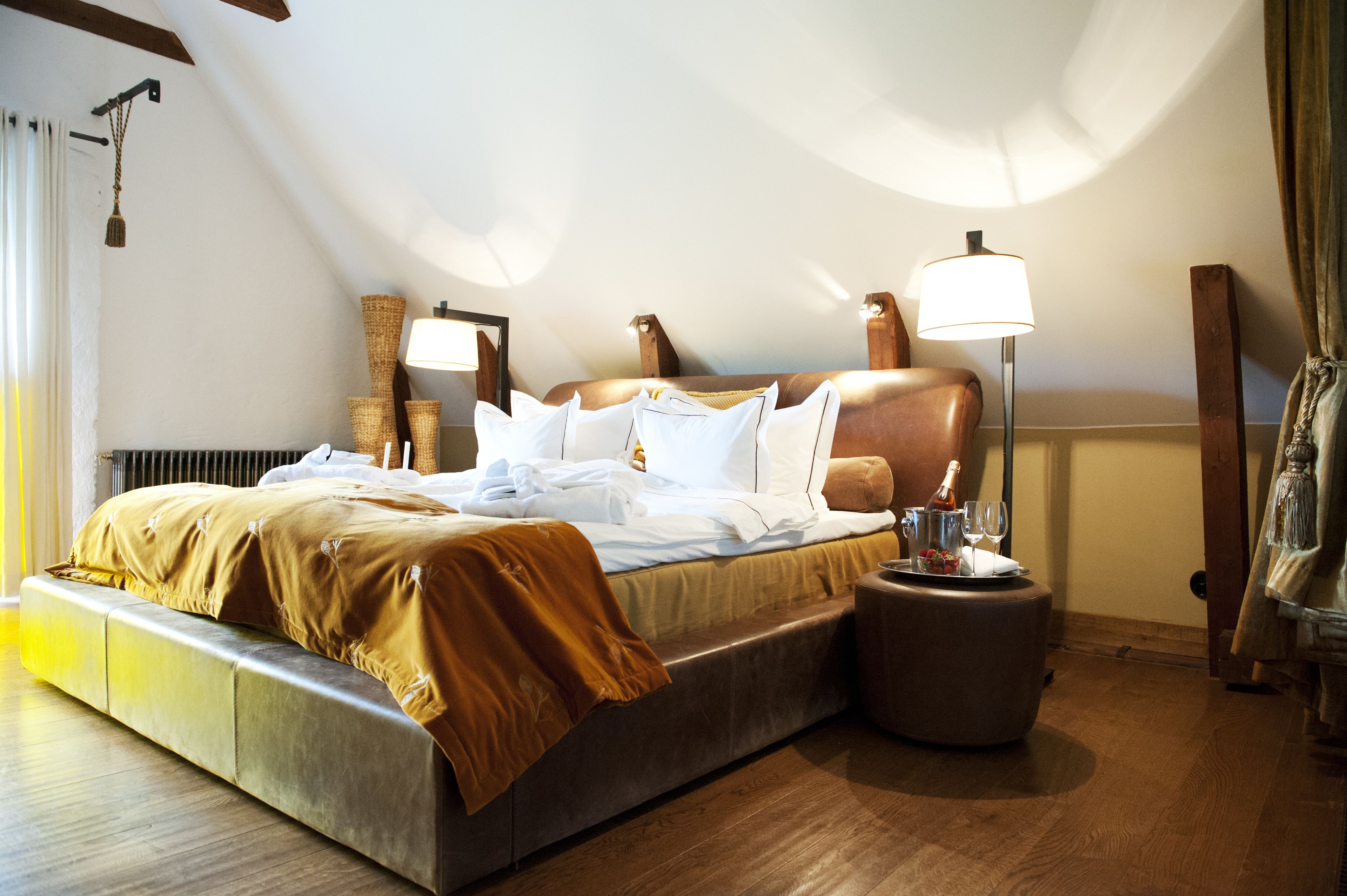 Una habitación de hotel moderna con una cama grande, dos lámparas de noche, una mesita redonda con una botella y vasos, y suelo de madera.