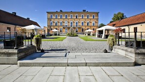 A courtyard with a central pathway leading to a large building with multiple windows and a tiled roof. There are outdoor seating areas with umbrellas on either side of the pathway. - Ulfsunda Slott (Bromma)