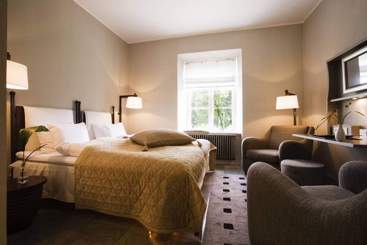 A hotel room with a bed, two armchairs, a side table, a lamp, and a window with a view of greenery.