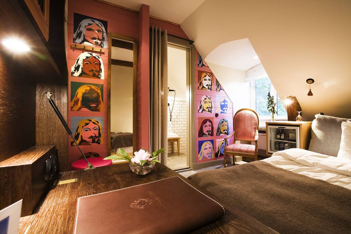 A bedroom with a bed, a bedside table, a chair, a small table with a vase, and a wall with colorful artwork.