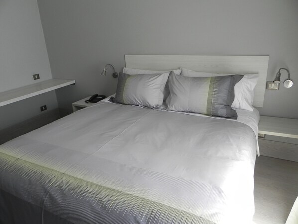 Una cama bien hecha con ropa de cama blanca y un almohadón verde y gris. Hay una mesita de noche con una lámpara, un teléfono y un pequeño reloj. La habitación tiene un diseño minimalista con paredes de gris claro.
