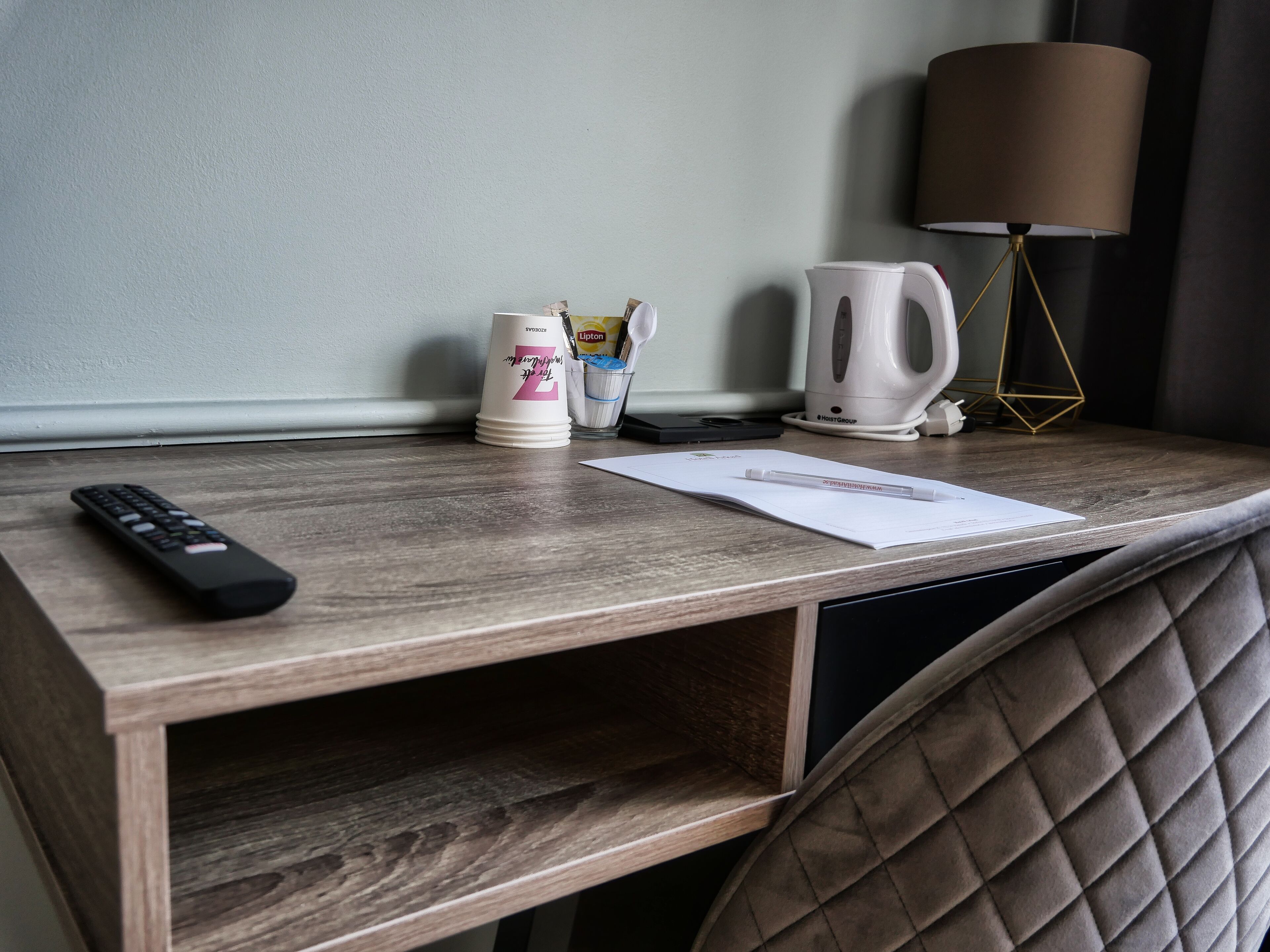 A wooden desk with a remote control, a white mug, a coffee maker, and a lamp.