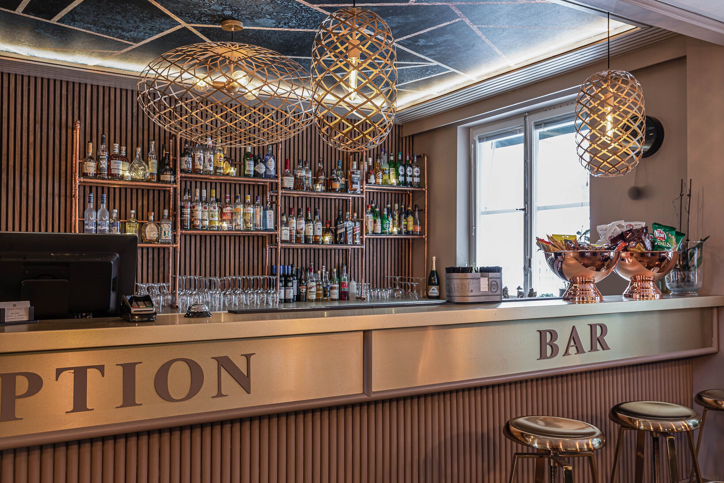 Zona de bar con un panel de madera, estantes con botellas y lámparas colgantes.