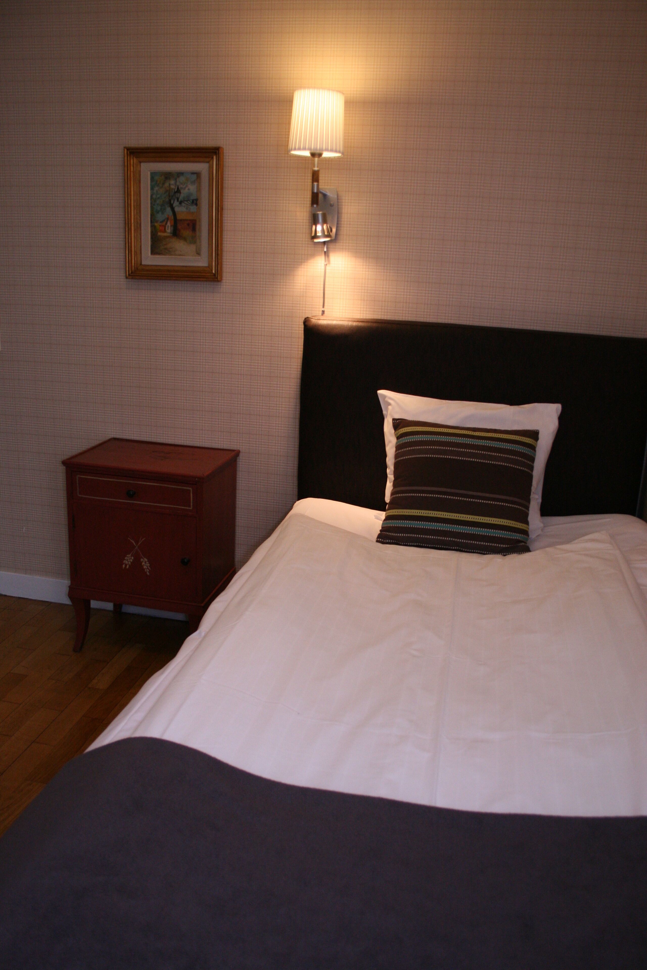 Una cama bien hecha con un cojín, una mesita de noche y una lámpara de pared.